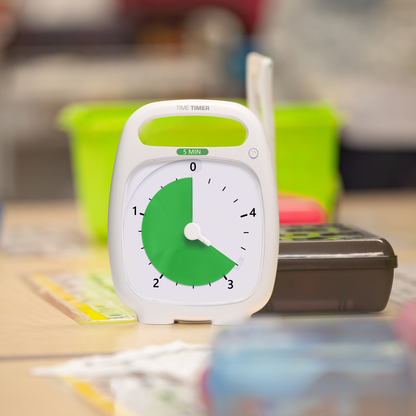 The Time Timer PLUS 5 minute visual timer is placed on a classroom desk. It is sitting in front of a pencil box and other school supplies. The green disk of the timer is set to 3 minutes and 15 seconds.