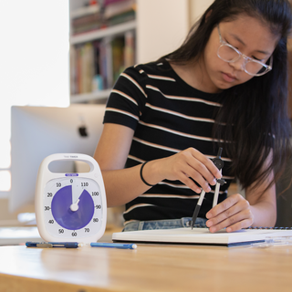 The Time Timer PLUS 120 Minute visual timer is placed in an art classroom on the drawing table. A young woman is in the background using a compass to draw in a sketchbook. The 2 hour visual timer is set to 118 minutes to show how much time is left in her studio class.