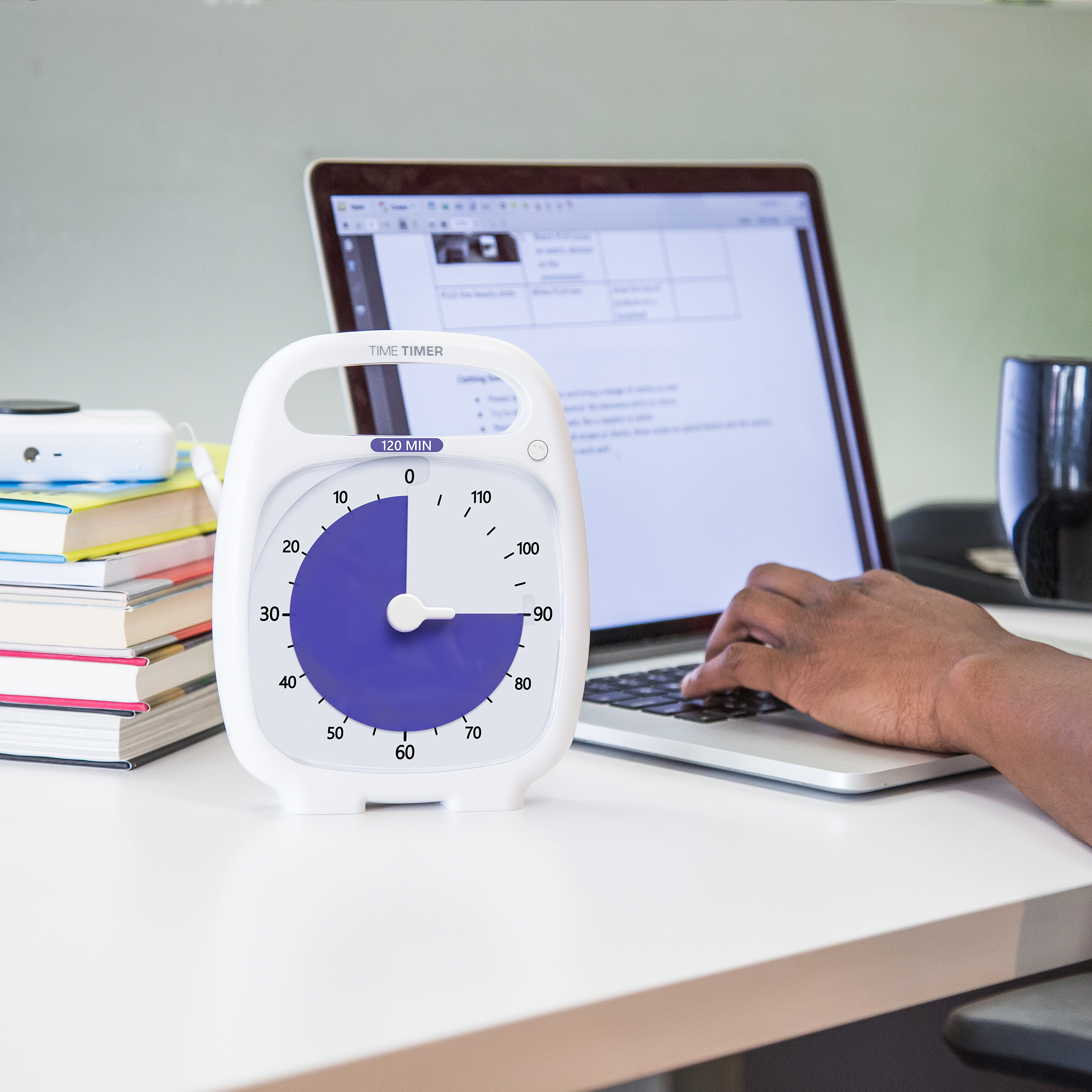 The Timer Timer PLUS 120 Minute visual timer is sitting on a desk in an office or home setting. Someone is using a laptop in the background and there is a pile of books sitting next to the computer. The purple disk of the Time Timer visual timer is set at 90 minutes to indicate a focused work or study session.
