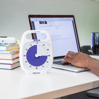 The Timer Timer PLUS 120 Minute visual timer is sitting on a desk in an office or home setting. Someone is using a laptop in the background and there is a pile of books sitting next to the computer. The purple disk of the Time Timer visual timer is set at 90 minutes to indicate a focused work or study session.