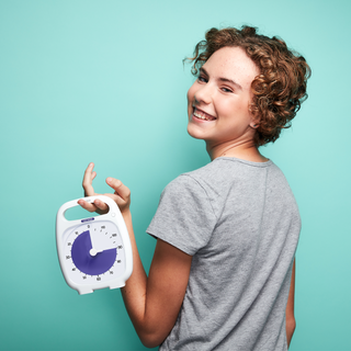 A smiling young woman with curly hair is standing in front of a bright blue background. She is holding the Time Timer 120 Minute timer. The purple disk is set to a 90 minute duration.