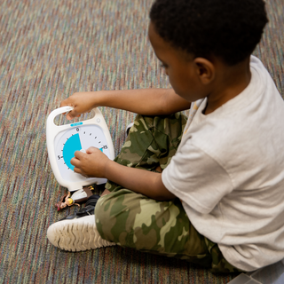 A young boy sits on the floor next to a small doll. He is setting the Time Timer PLUS 20 Minute Visual Timer by using the center knob. He is turning the duration to about 15 minutes.