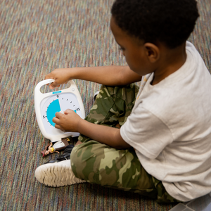 A young boy sits on the floor next to a small doll. He is setting the Time Timer PLUS 20 Minute Visual Timer by using the center knob. He is turning the duration to about 15 minutes.