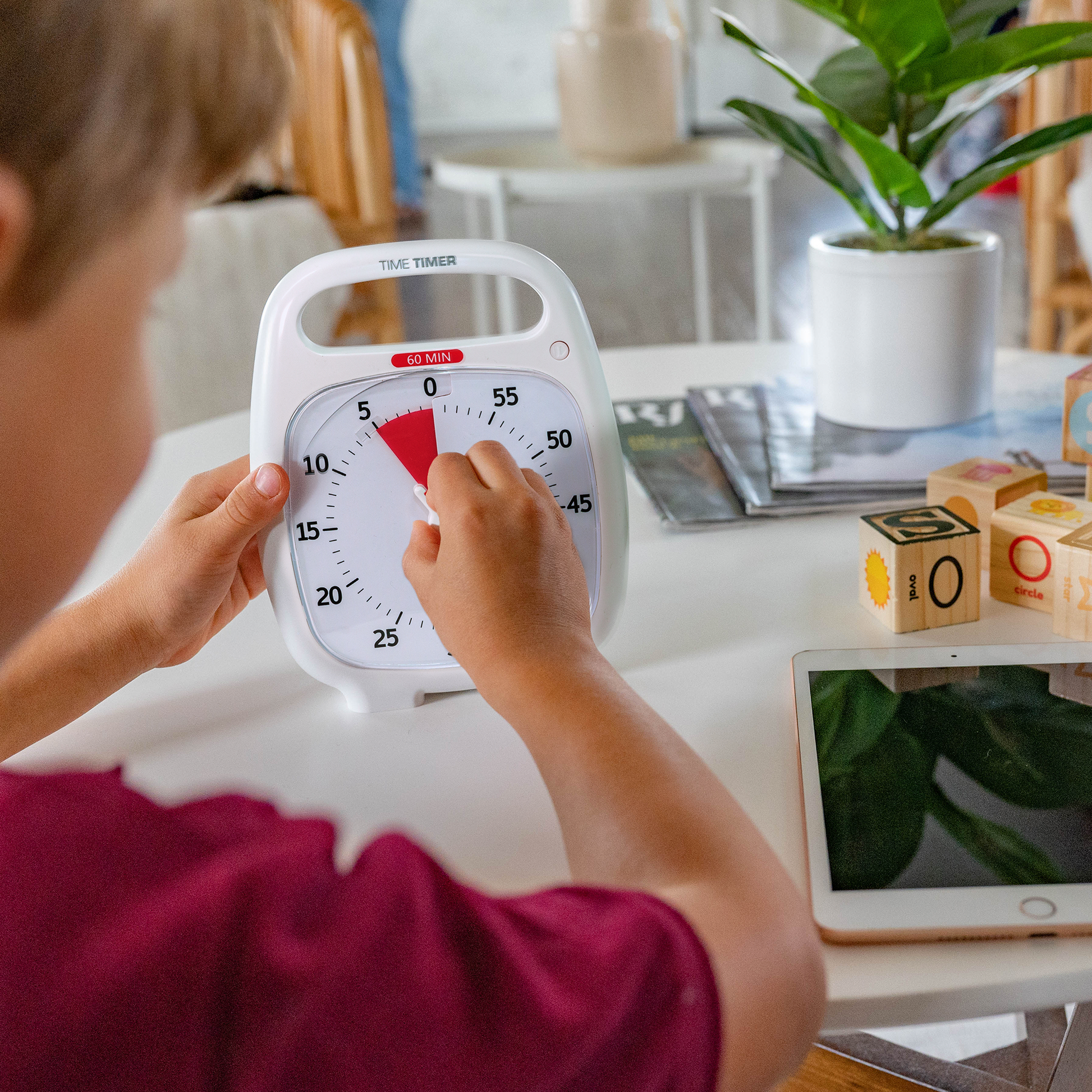 A young boy is setting the Time Timer PLUS 60 Minute visual timer in white. He is turning the knob to set the red disk to 5 minutes. There is a tablet and blocks on the table.