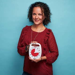 A smiling woman is standing in front of a blue background and holding the Time Timer PLUS 60 minute visual timer in white. The red disk of the timer is set to 45 Minutes.