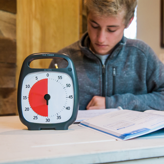 A young man is sitting at a table doing homework. The Time Timer PLUS 60 minute visual timer in the charcoal color is sitting in front of him. The red disk on the timer is set to 30 minutes.